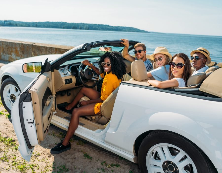 multicultural young friends sitting in car and looking at camera at seaside